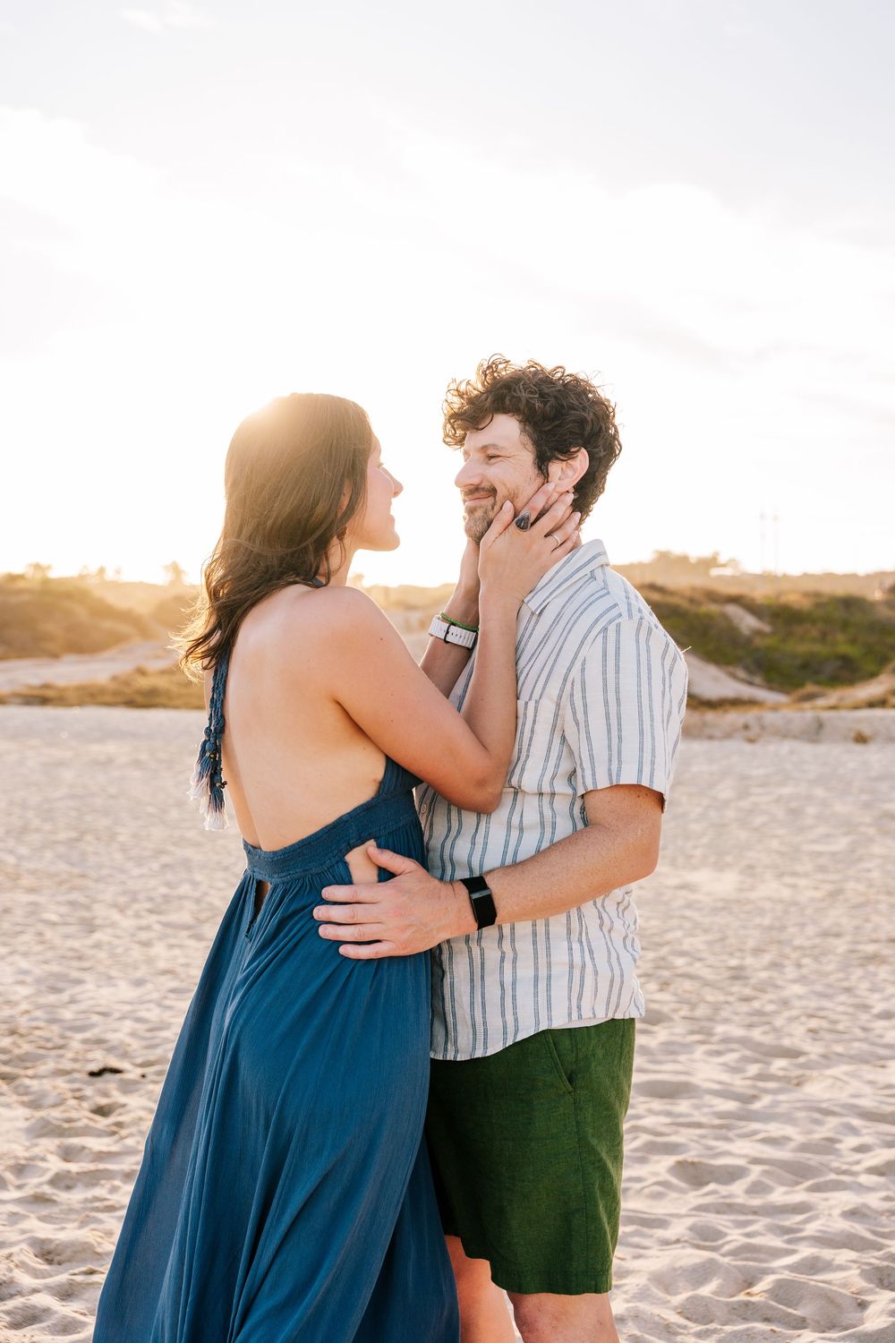 Couple embracing on sandy beach at golden hour, woman in blue backless dress touching man's face.