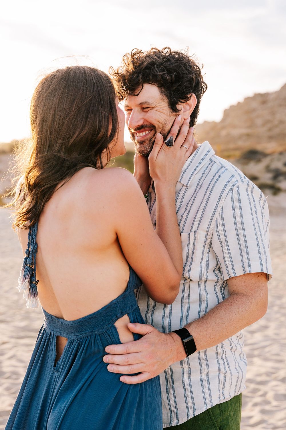 A couple shares a tender moment on a sandy beach at golden hour, woman in blue dress touching mans face.