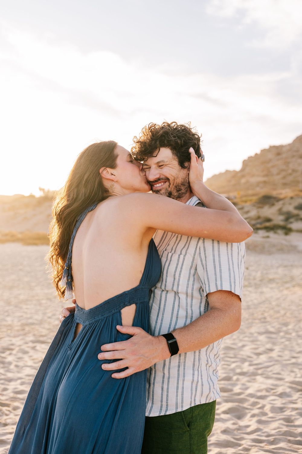 Woman in blue backless dress kissing man's cheek while embracing on beach at sunset.