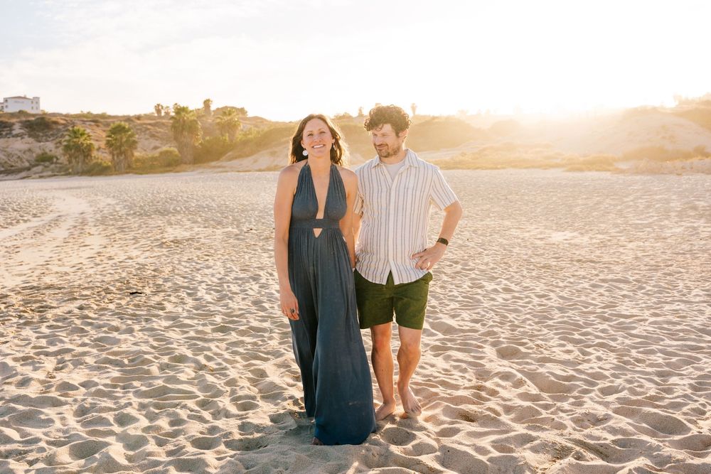 Couple walking on sandy beach at golden sunset, woman in navy maxi dress, man in striped shirt and green shorts.