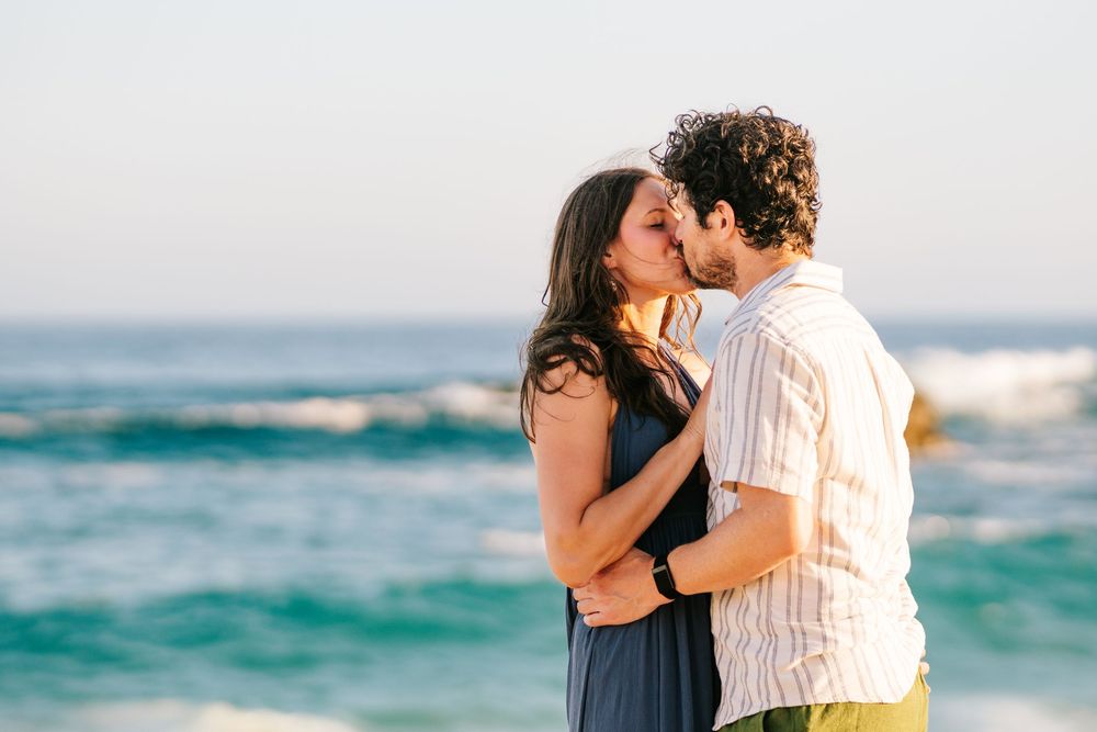 A couple shares a romantic kiss on a sunny beach with turquoise ocean waves in the background.