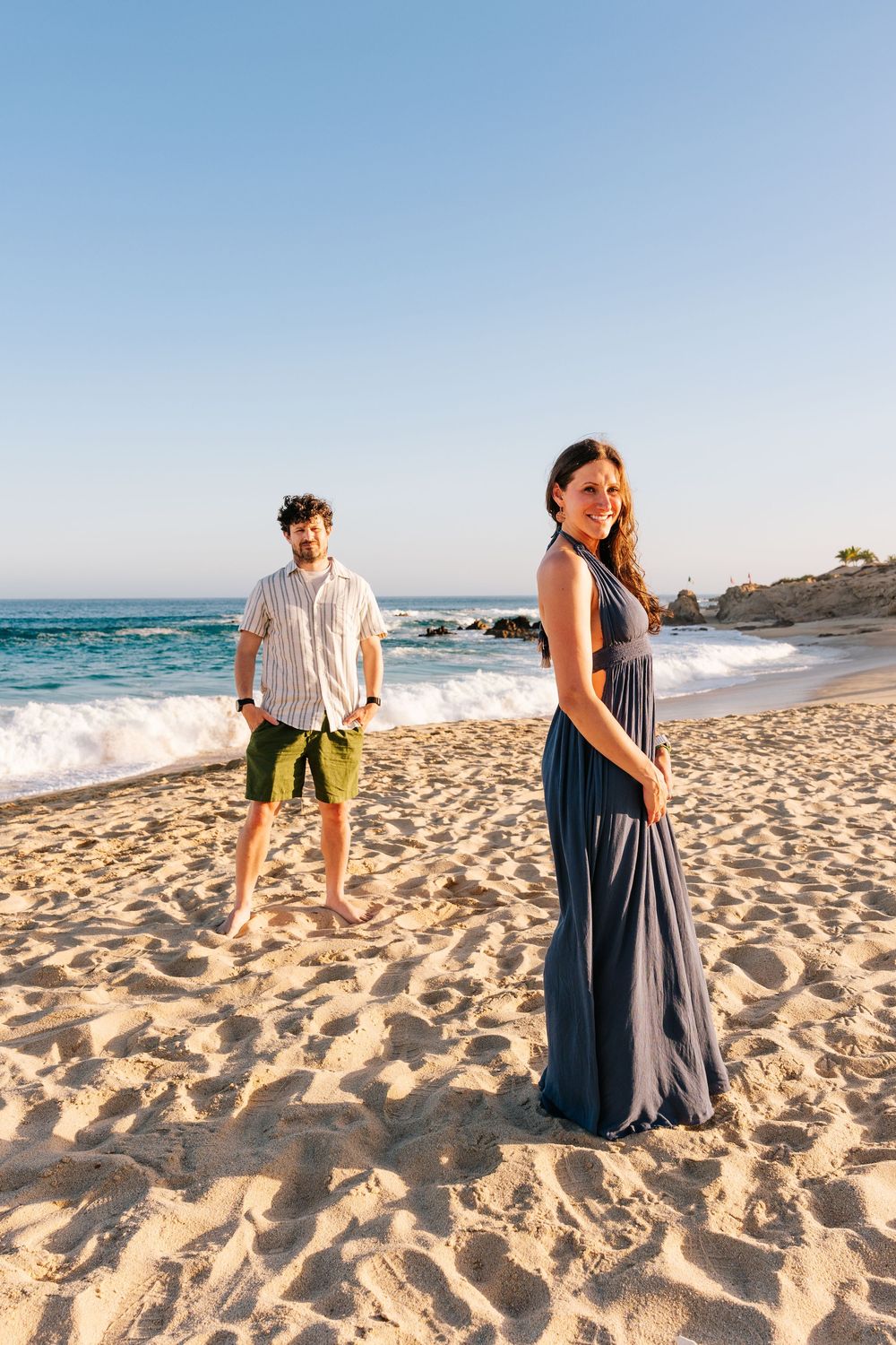 A couple poses on a sandy beach, woman in blue maxi dress smiling, man in green shorts stands behind near ocean waves.