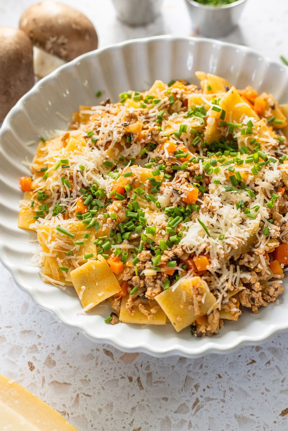A white bowl filled with pappardelle pasta, ground meat, carrots, topped with grated Parmesan and fresh chives.