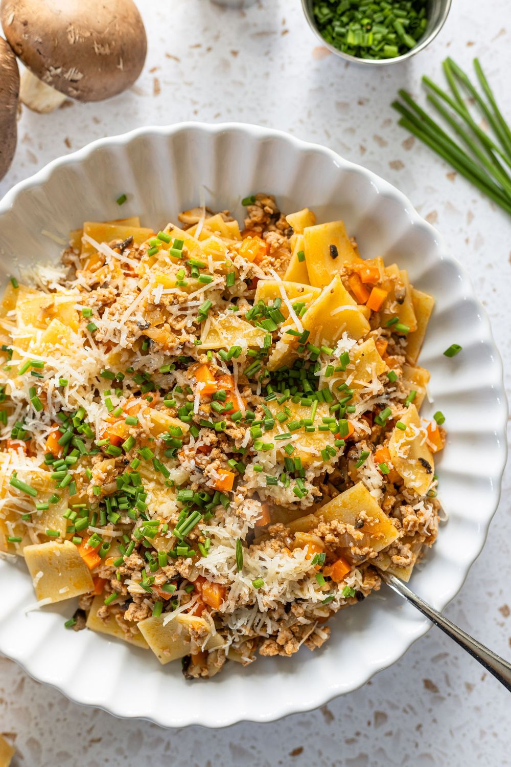 A bowl of pappardelle pasta with mushroom meat sauce, grated parmesan, and fresh chives on a white plate.