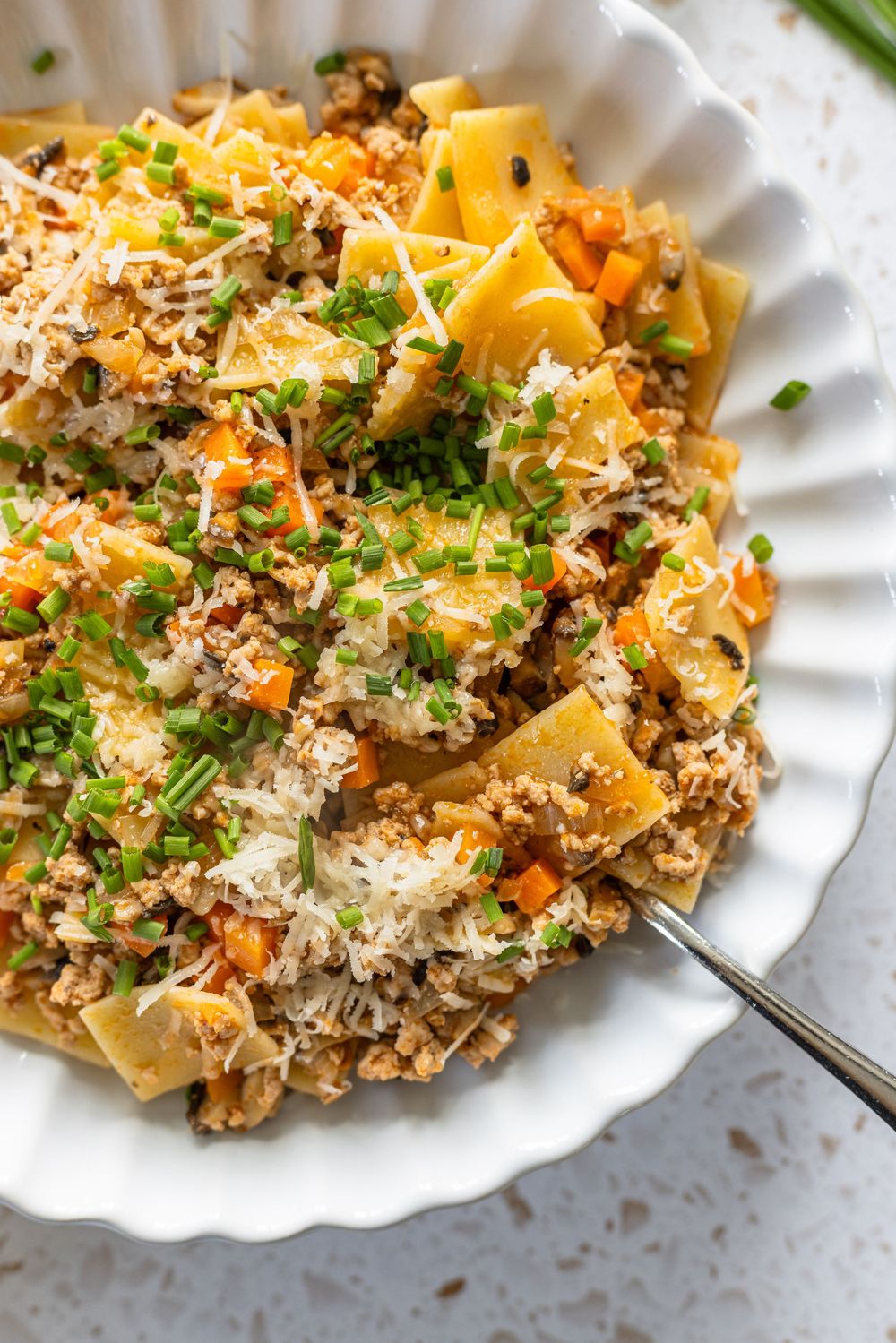 White bowl of pappardelle pasta with meat sauce, carrots, Parmesan, and fresh chives on a light surface.
