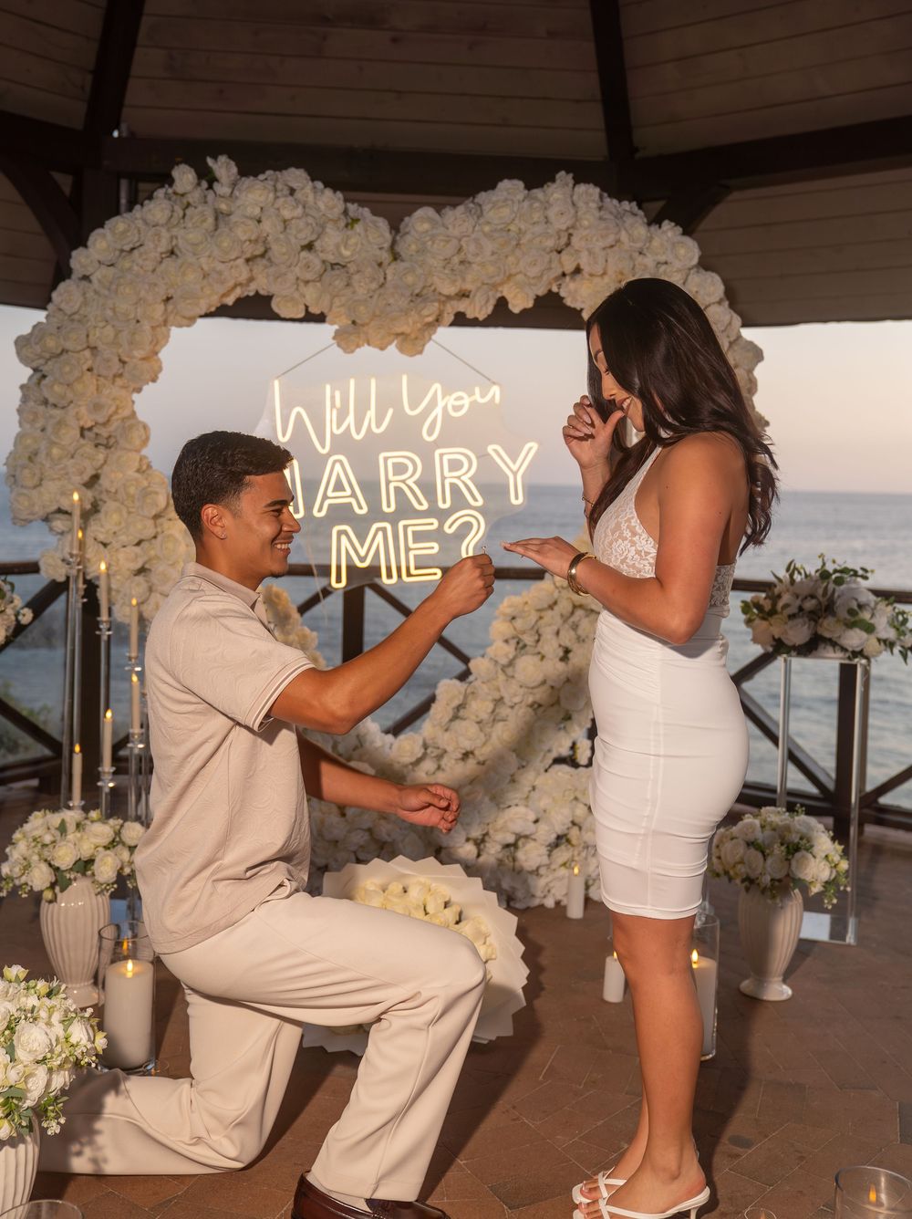 Man proposes to woman on one knee in front of white floral heart arch with Will You Marry Me neon sign.