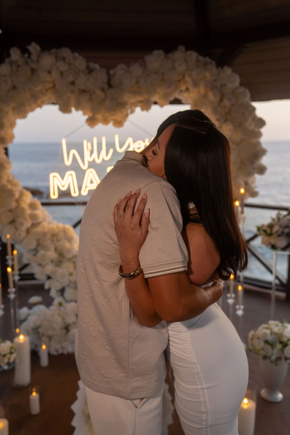 Couple embracing during a romantic marriage proposal with floral arch, candles, and neon sign by the ocean at dusk.