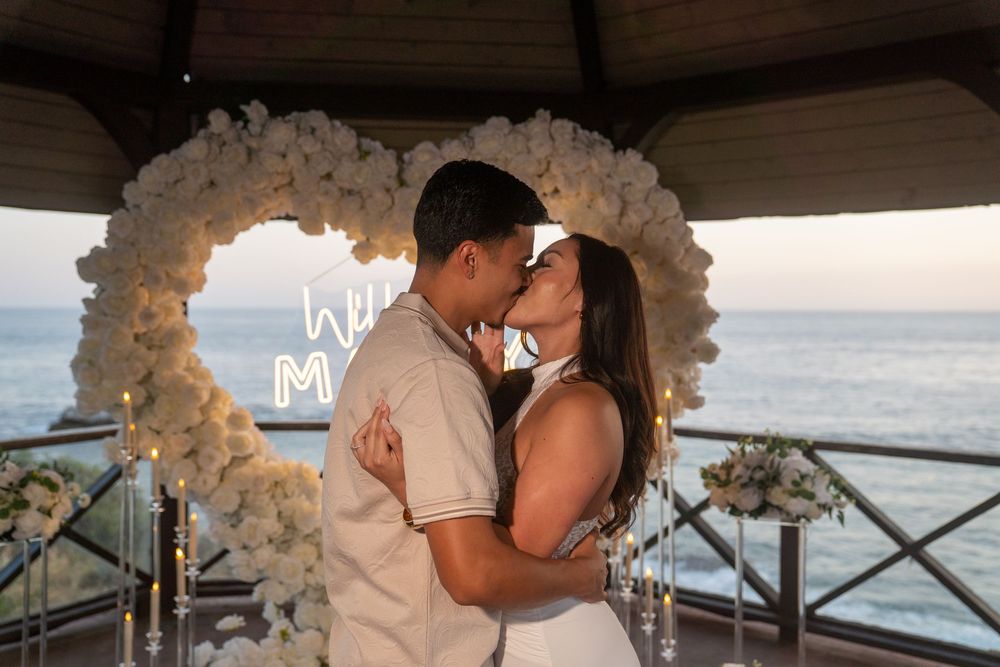 Couple sharing a romantic kiss under a white floral heart arch with ocean view at sunset proposal.