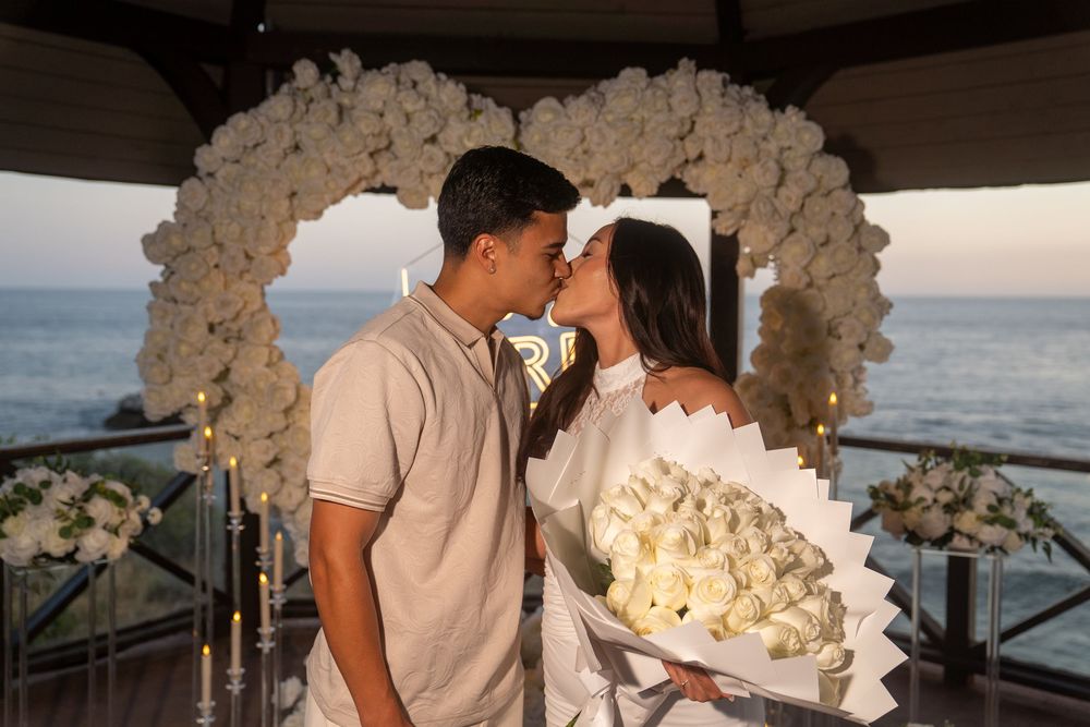 Couple kissing in front of white heart floral arch by the ocean, holding large white rose bouquet at romantic proposal.
