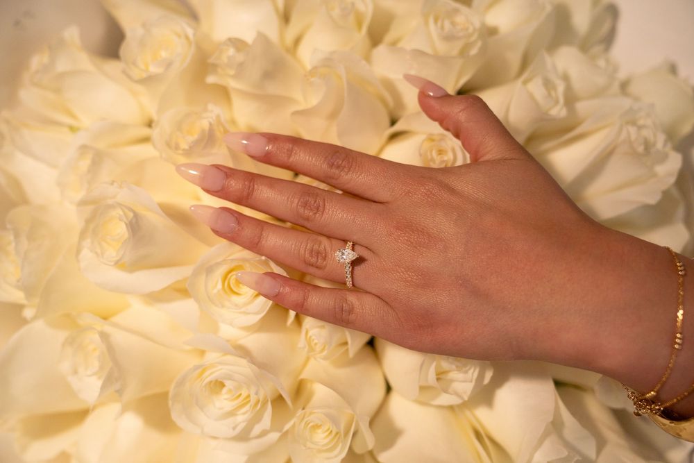 A hand with long nails wearing a diamond engagement ring resting on white roses.