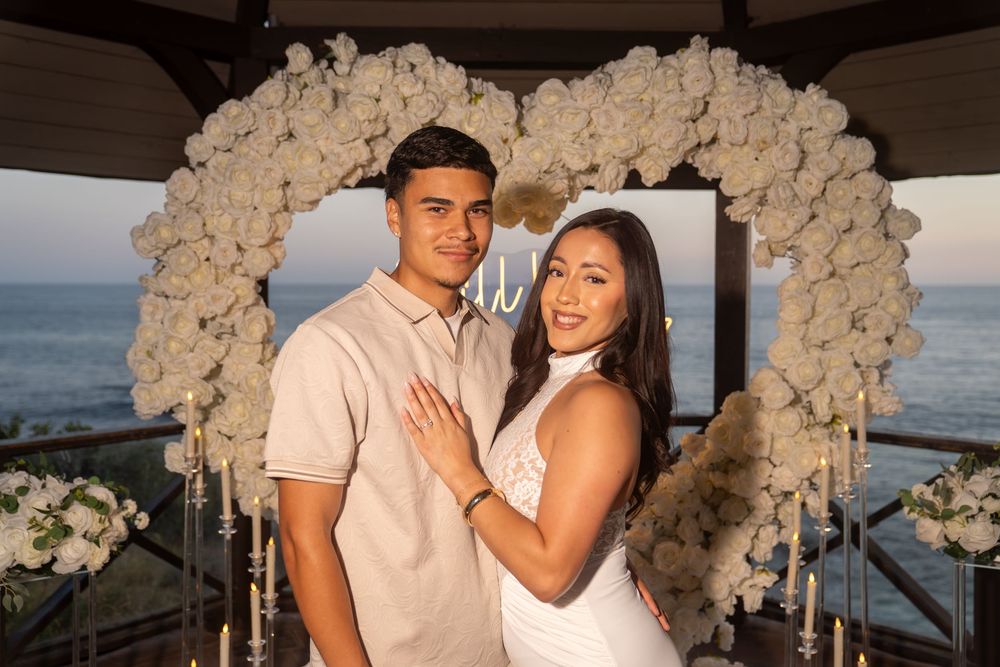 Couple poses in front of a white floral heart arch by the ocean at an engagement proposal setting.