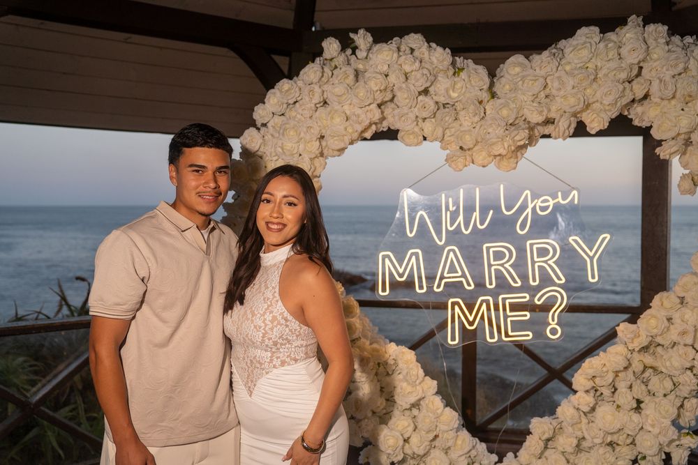 Couple smiling in front of a floral heart arch with a Will You Marry Me neon sign by the ocean.