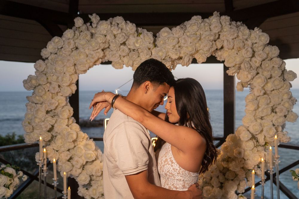 Couple embraces under a white floral heart arch with candles and ocean view at a romantic proposal setting.