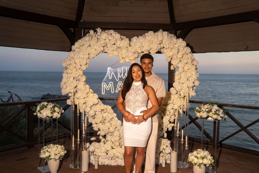Couple poses in front of a white floral heart arch with a neon sign by the ocean at a romantic proposal setup.