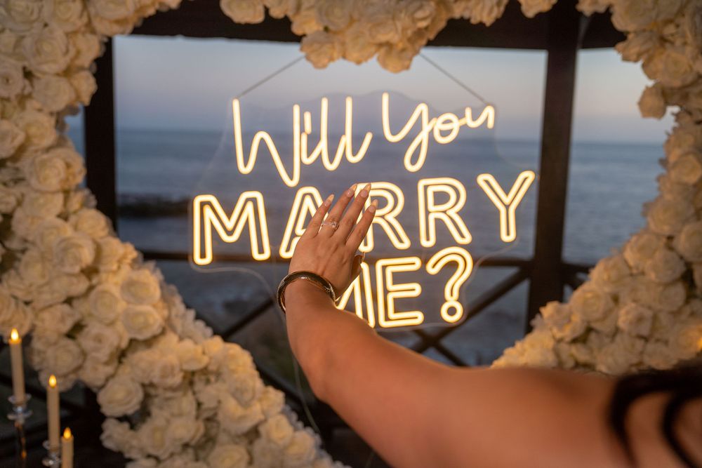Romantic marriage proposal neon sign saying Will You Marry Me surrounded by white flowers and candles at sunset.