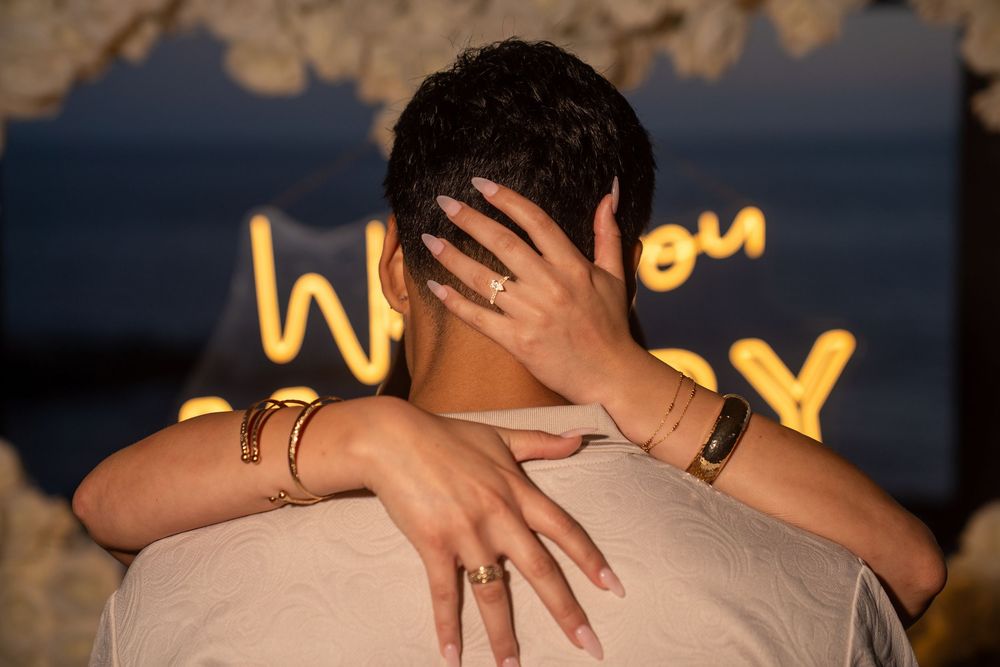 Couple embracing during romantic engagement proposal with glowing neon sign in background at sunset.