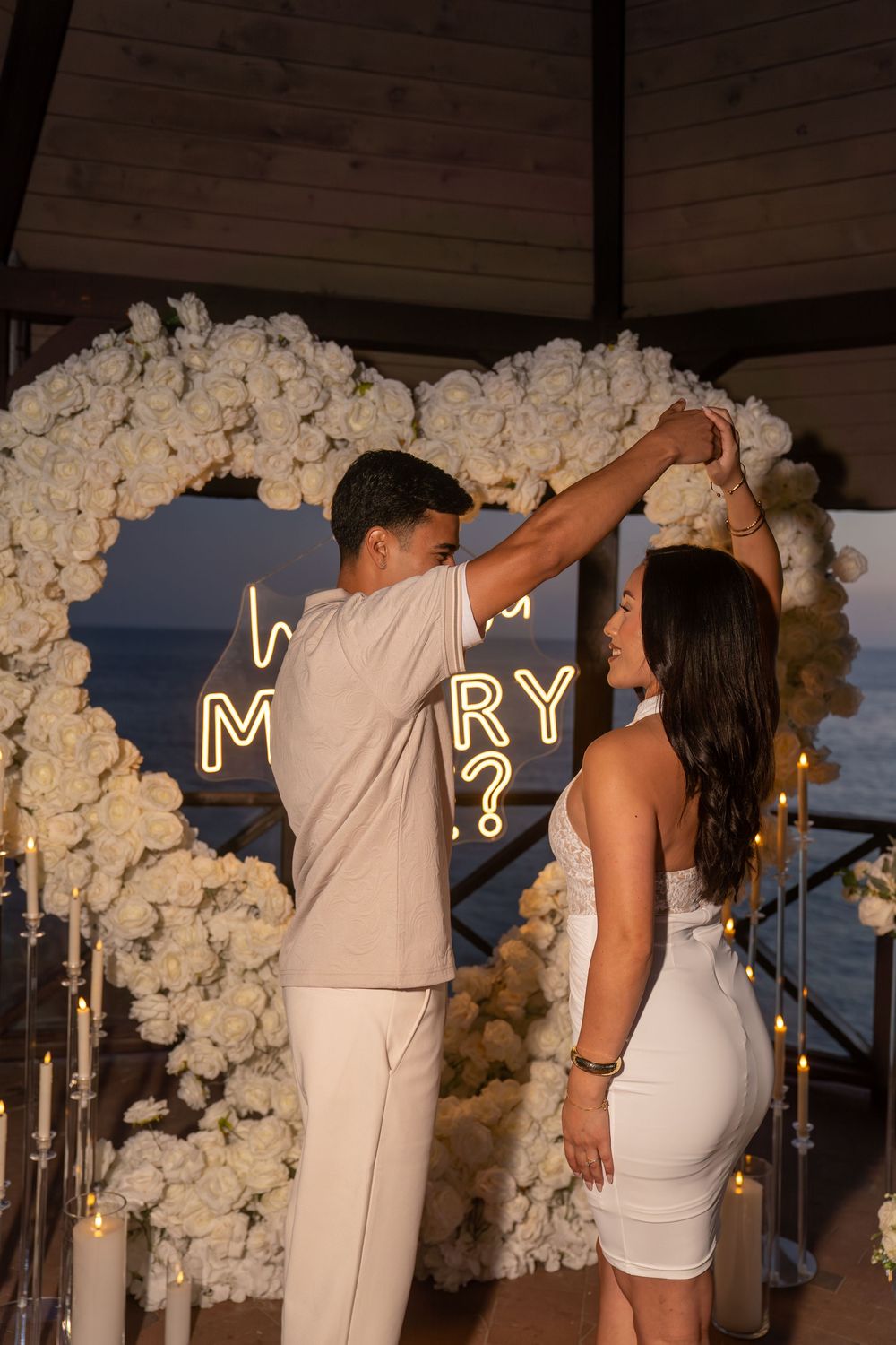 Couple dancing under a white floral heart arch with a "Will You Marry Me?" neon sign at a seaside proposal.