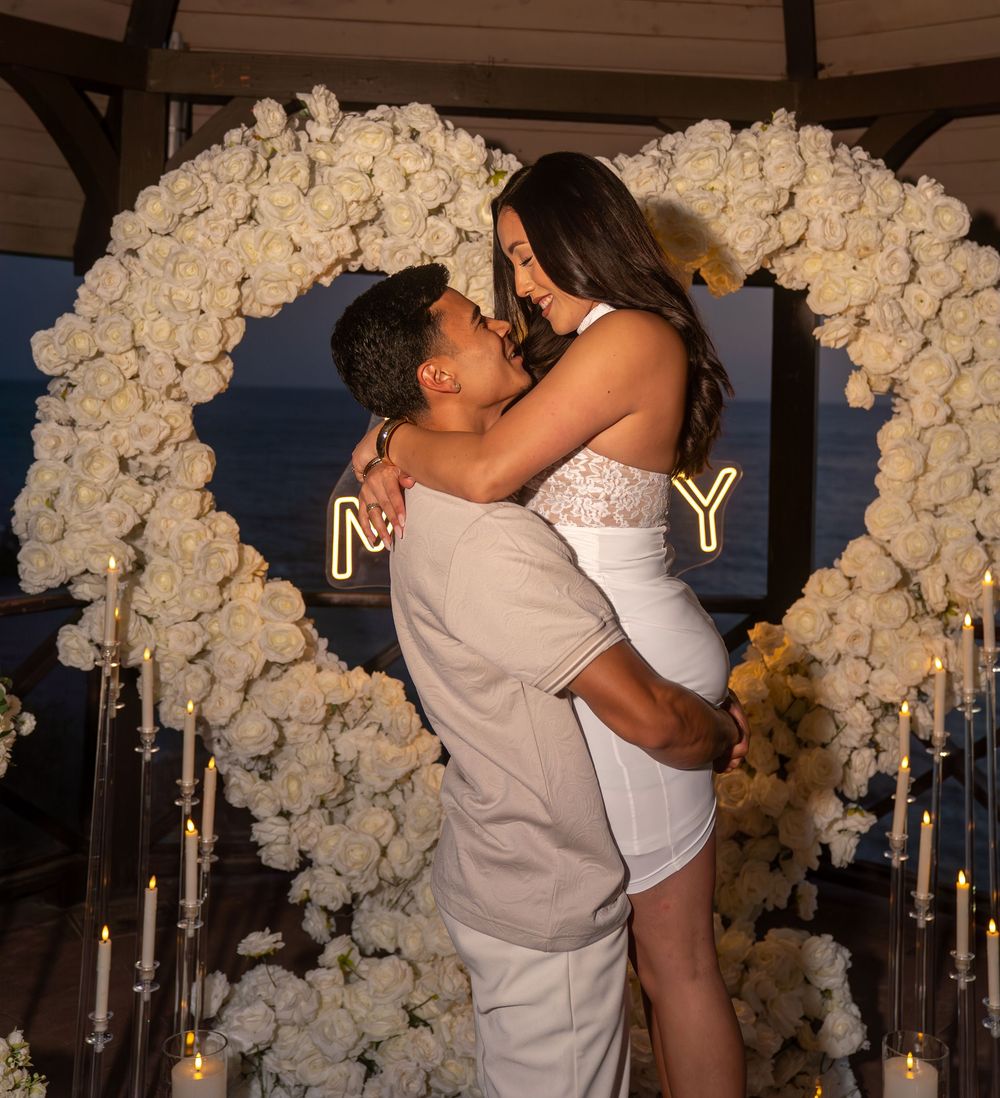 Couple embracing in front of a white rose heart arch with candles during a romantic beach proposal.