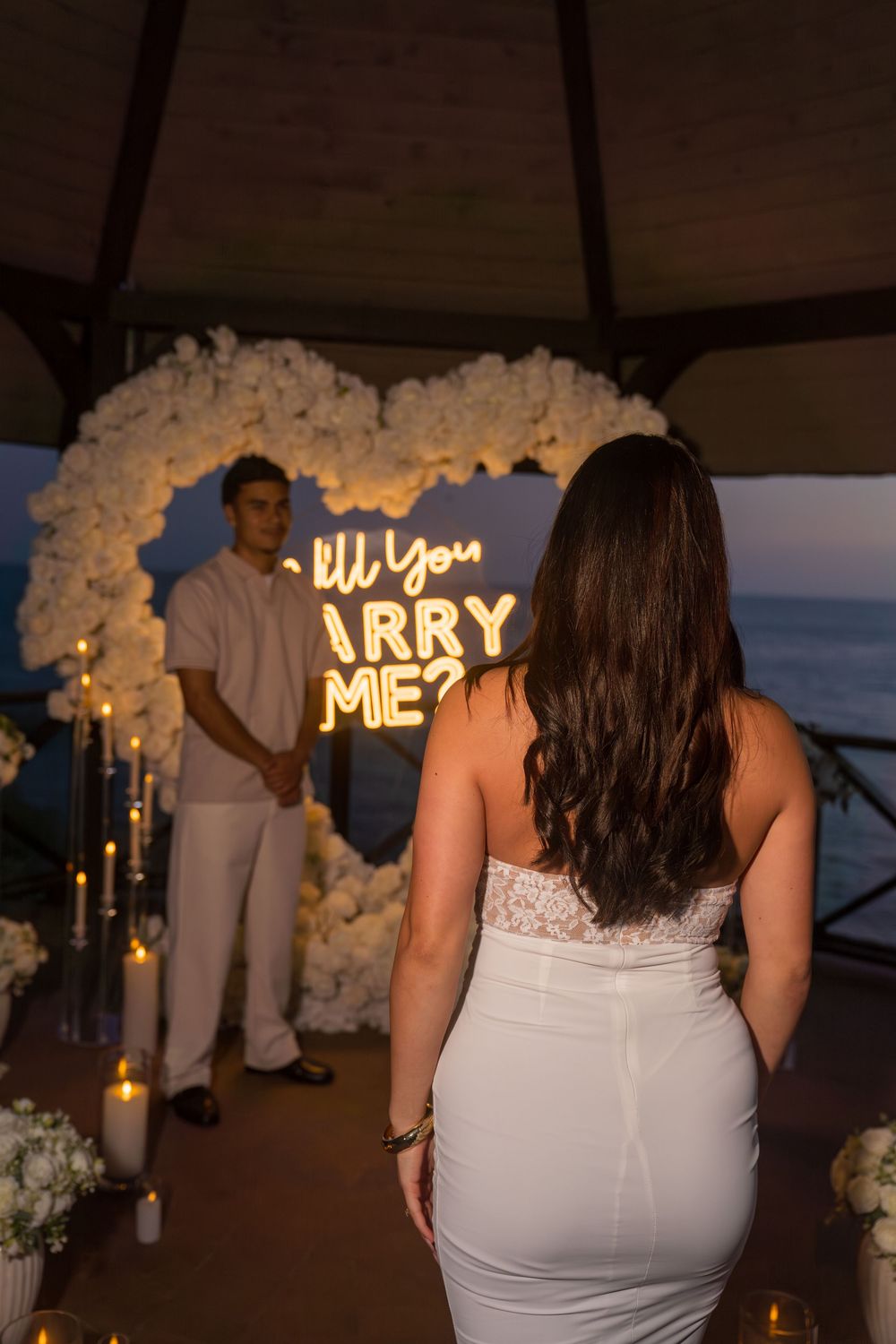 Woman in white dress approaches man standing before romantic floral heart arch with Will You Marry Me neon sign.