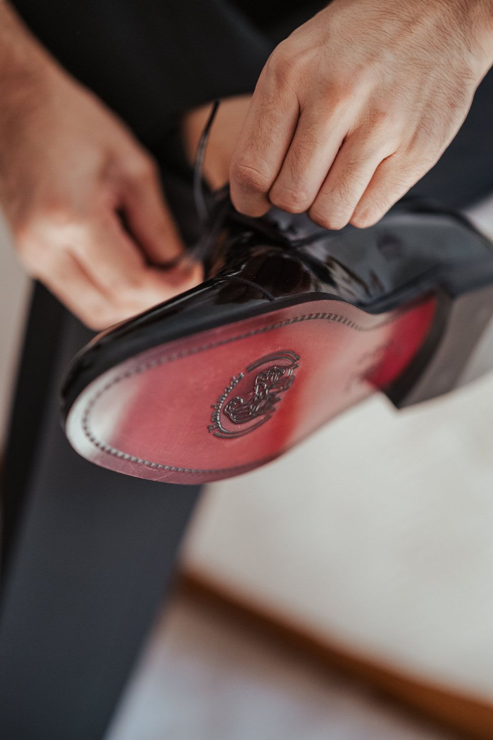 Hands tying laces on a black dress shoe with a red leather sole and embossed logo.