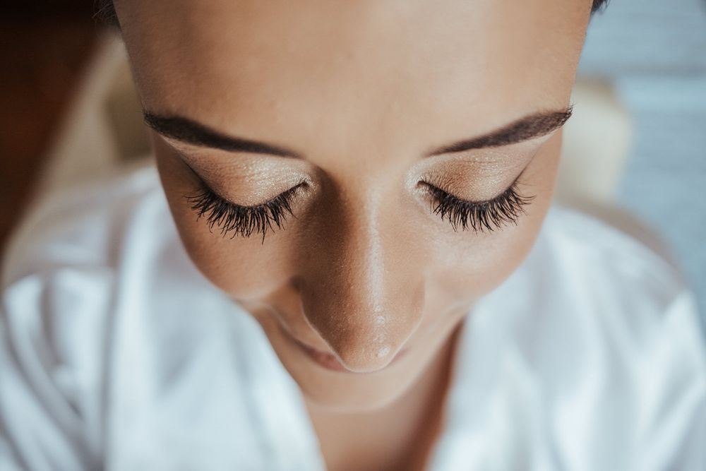 Close-up of a woman with long eyelashes and groomed eyebrows, eyes closed, wearing a white robe.