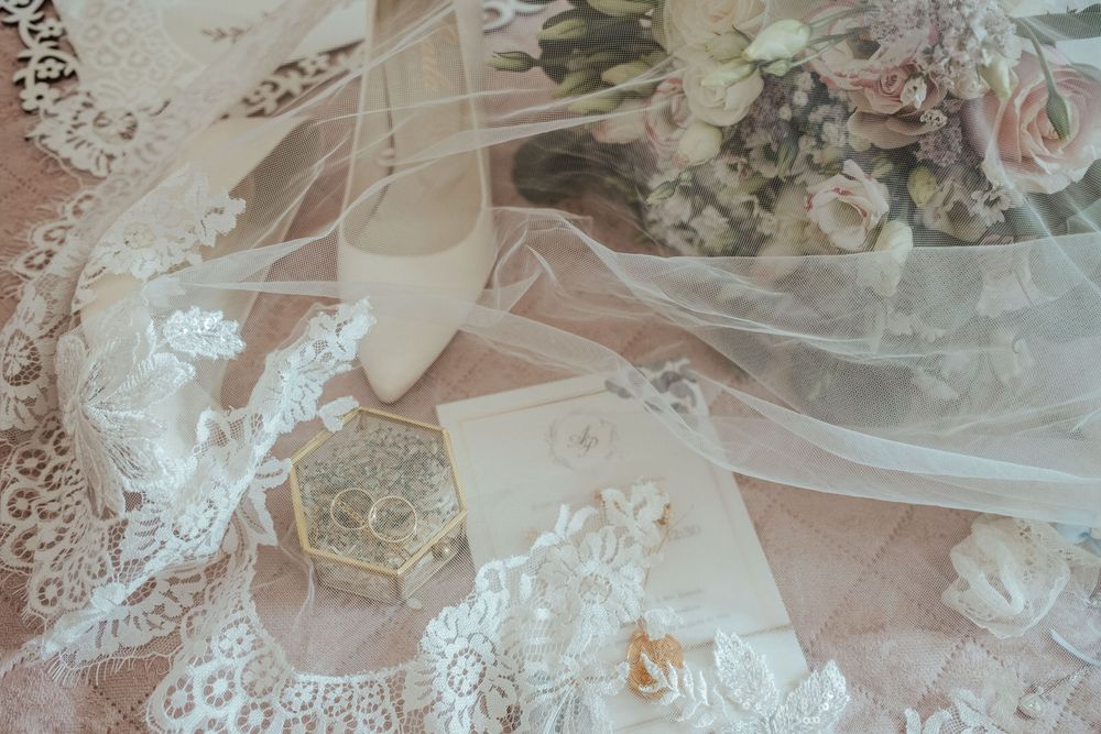 Elegant wedding flat lay with lace veil, bridal shoe, floral bouquet, gold ring box, and invitation cards.