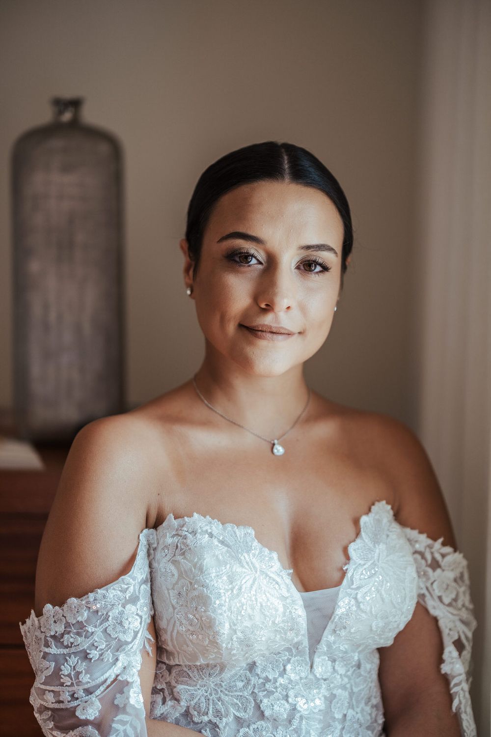 Bride in off-shoulder lace wedding gown with pearl necklace, elegant updo hairstyle, soft natural makeup, indoor portrait.