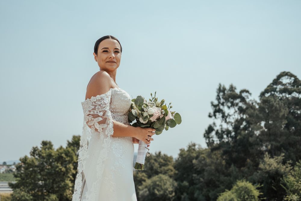 Bride in lace off-shoulder wedding dress holding white floral bouquet, posing outdoors with trees and blue sky.