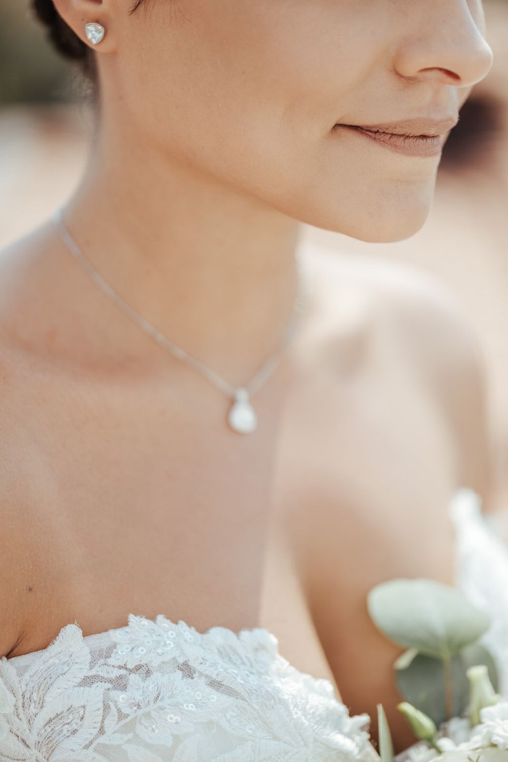 Bride wearing a pearl drop necklace and stud earrings with a white lace strapless gown, holding a bouquet.