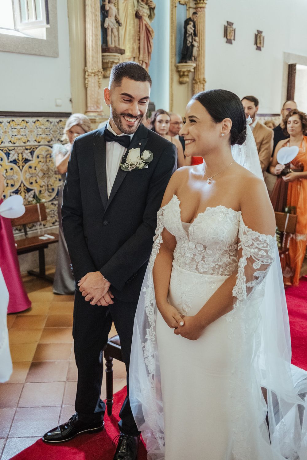 Bride and groom smiling at each other during wedding ceremony in a church, bride in lace off-shoulder gown.