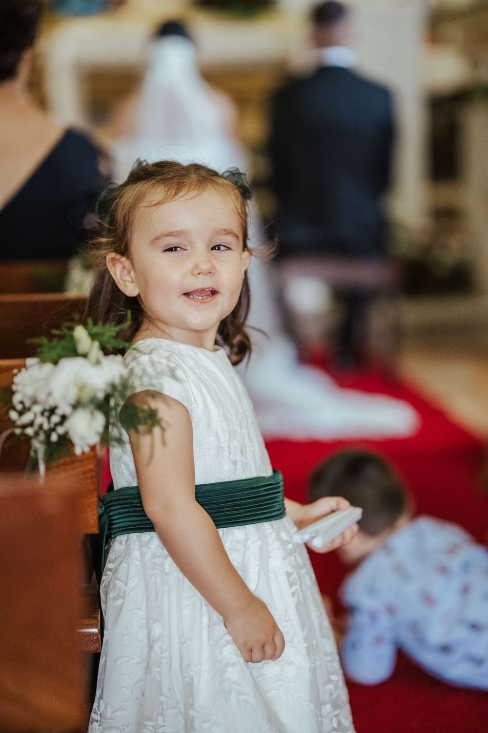 A smiling flower girl in a white dress with green sash stands in a church aisle during a wedding ceremony.