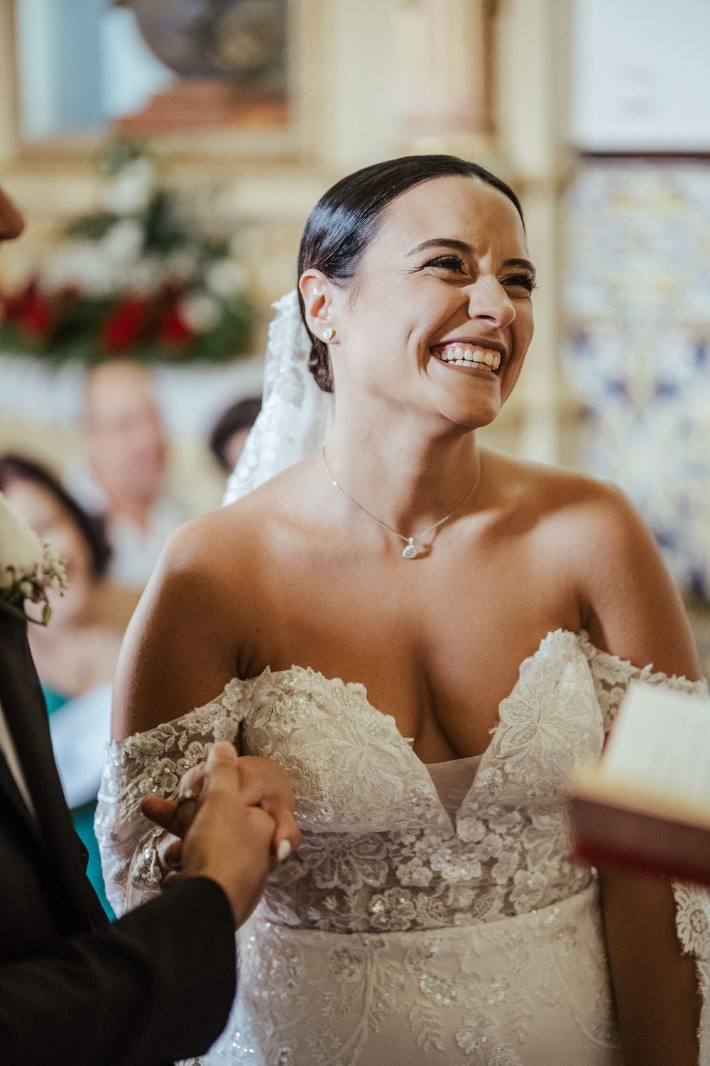 Radiant bride in lace off-shoulder gown laughing joyfully during wedding ceremony while holding hands at the altar.