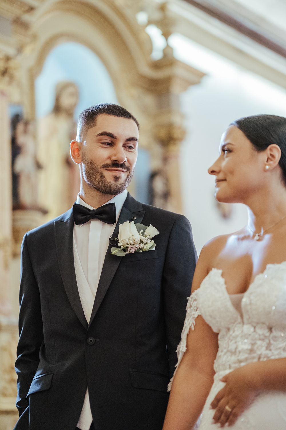 Groom in black tuxedo with boutonniere smiles at bride in white lace gown during church wedding ceremony.