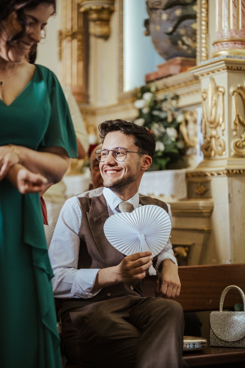 Smiling man in glasses and brown vest holds a white paper fan while sitting in an ornate church during a wedding.