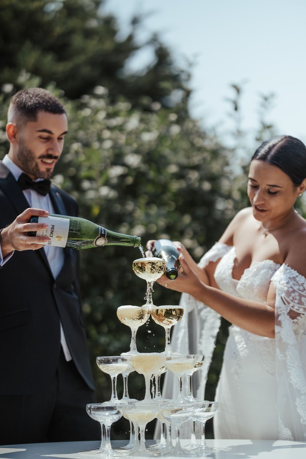 Bride and groom pouring champagne tower at outdoor wedding celebration.