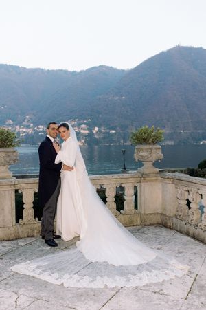 Bride and groom pose on elegant stone terrace overlooking Lake Como with mountain backdrop on their wedding day.