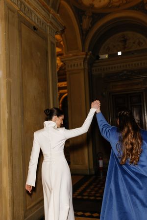 Two women in white and blue coats hold hands while walking through an ornate classical building interior.