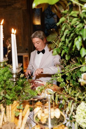 Elegant man in white tuxedo carving meat at a candlelit reception table adorned with lush greenery and floral arrangements.