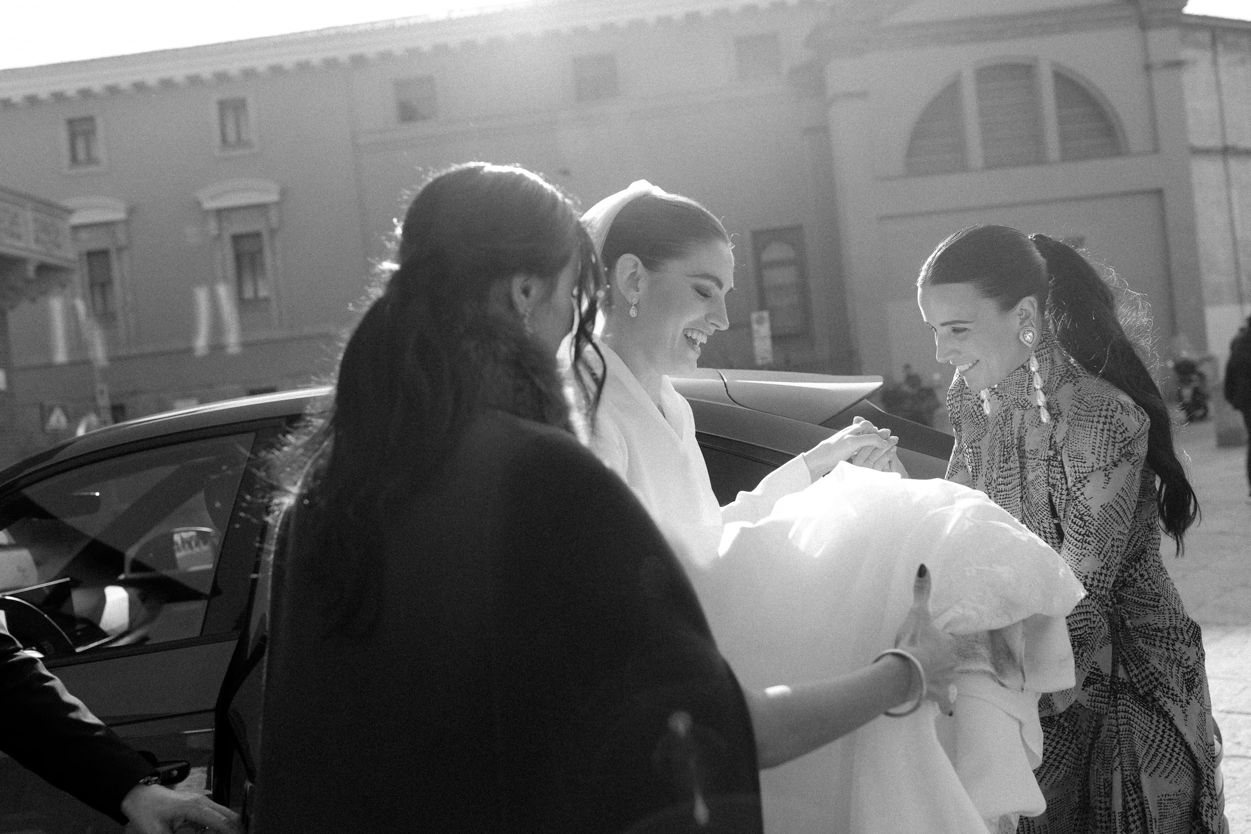Black and white photo of a bride being helped into a car by two women outside a historic building.