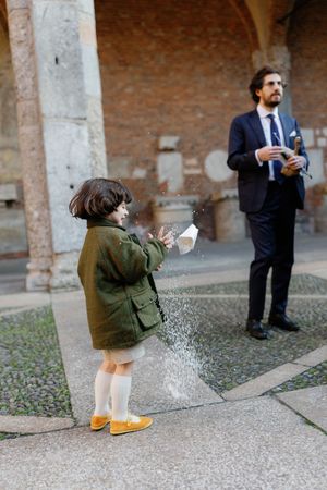 Child tosses rice confetti outside rustic brick venue as suited musician plays violin at wedding.