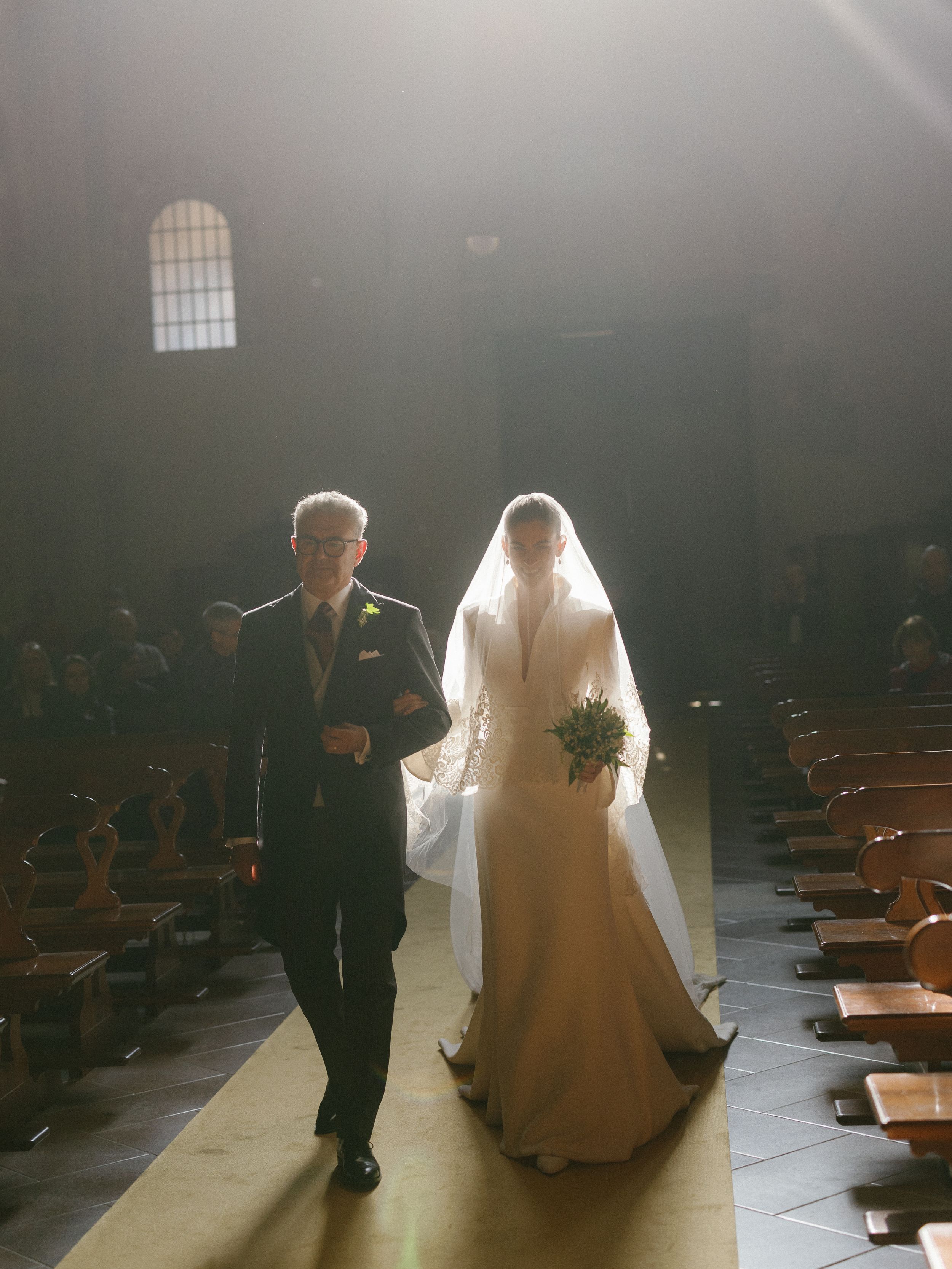 Bride in white gown and veil walks down sunlit church aisle with escort.
