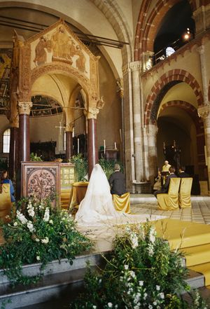 Bride in white veil kneels at altar in ornate Romanesque church decorated with yellow flowers.