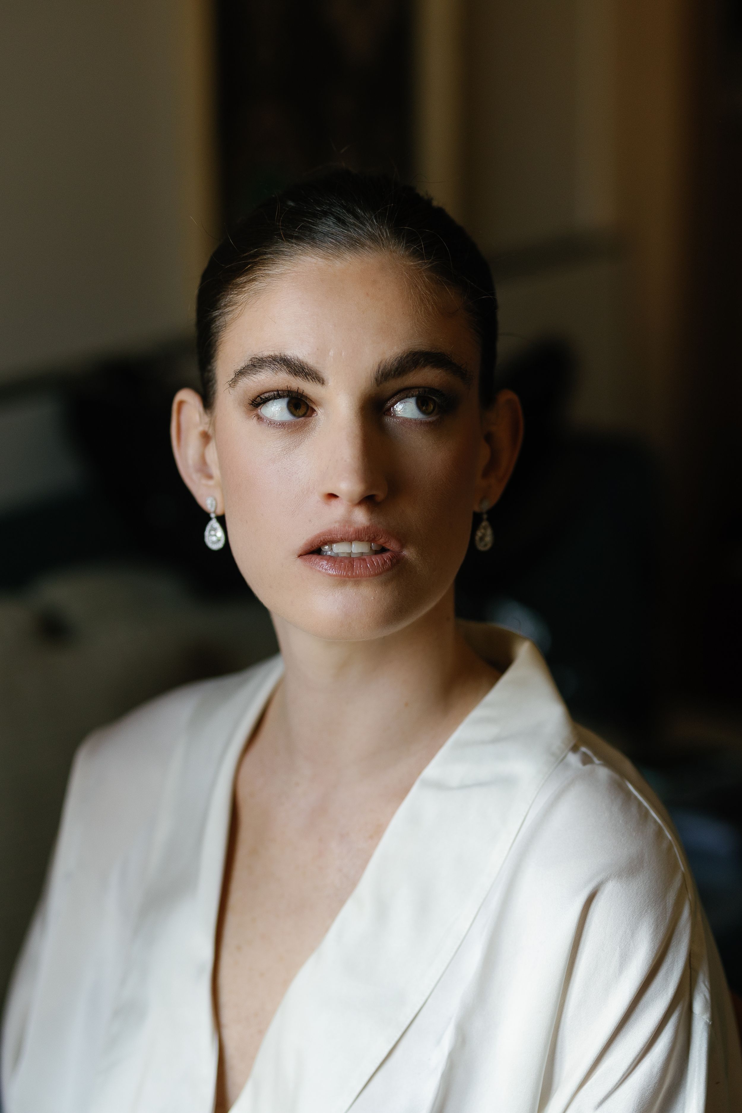Bride with sleek updo and drop earrings wearing white robe during wedding preparation.