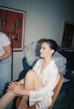 Bride in white silk robe sits in chair during wedding preparations, with art on walls in background.