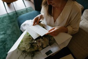 Woman in white blouse reading handwritten letter beside white floral bouquet on a couch.