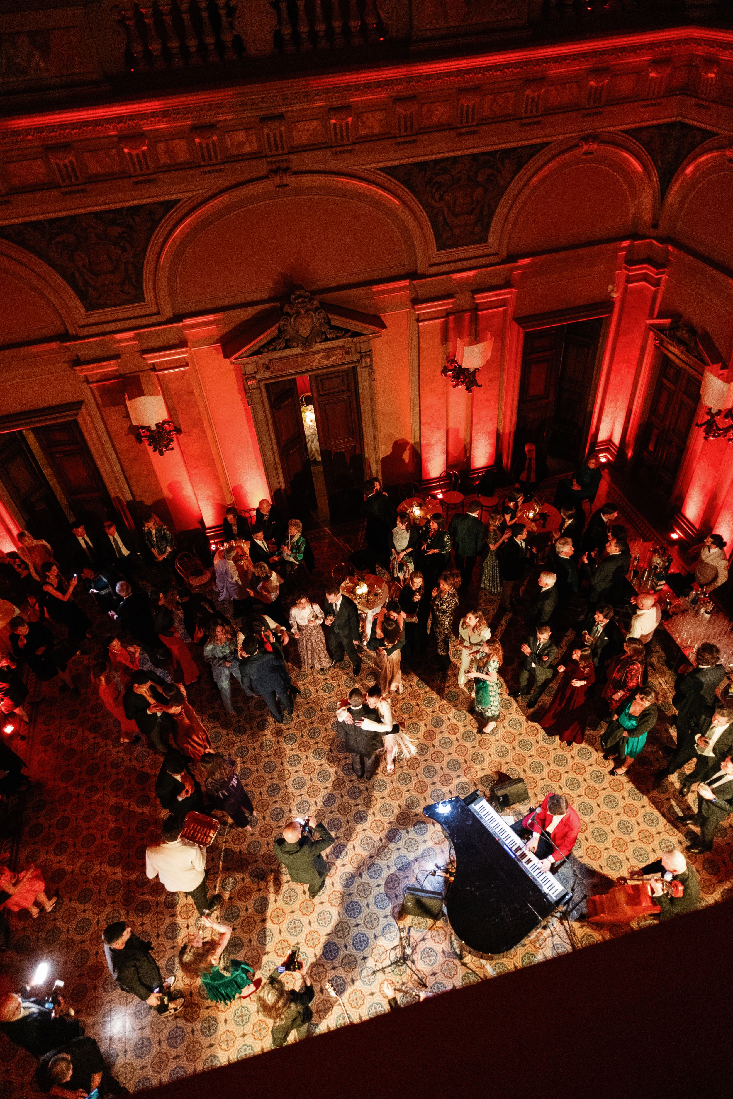 Aerial view of elegant ballroom event with red lighting, ornate architecture, and guests dancing on decorative tiled floor.