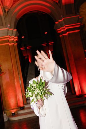 Bride in white gown holds bouquet of lily of the valley while blocking camera with hand in red-lit venue.