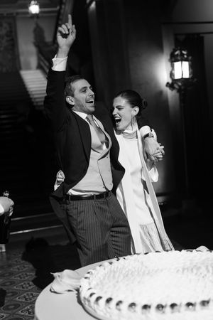 Black and white photo of joyful newlyweds celebrating beside their wedding cake, groom raising fist in triumph.