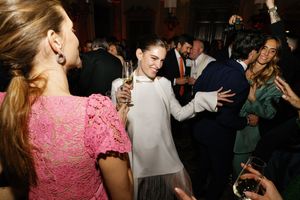 Guests in elegant attire laugh and dance at an upscale indoor party, woman in white holding champagne.