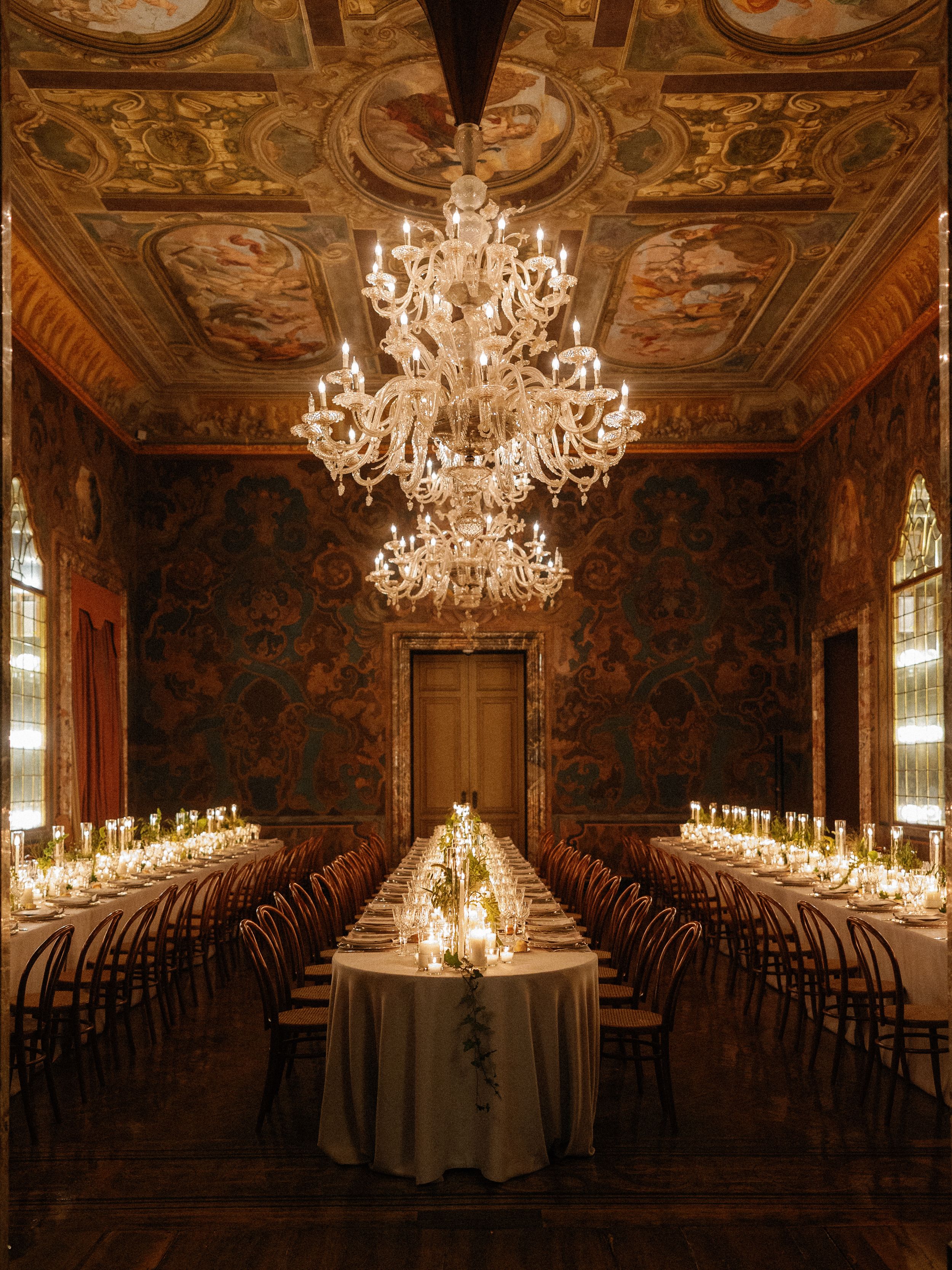 Opulent wedding reception hall with crystal chandelier and candlelit long dining tables.